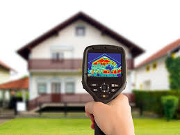 image of hand holding a thermal camera which is pointed at a house. The camera's display is showing a thermal image of the house.